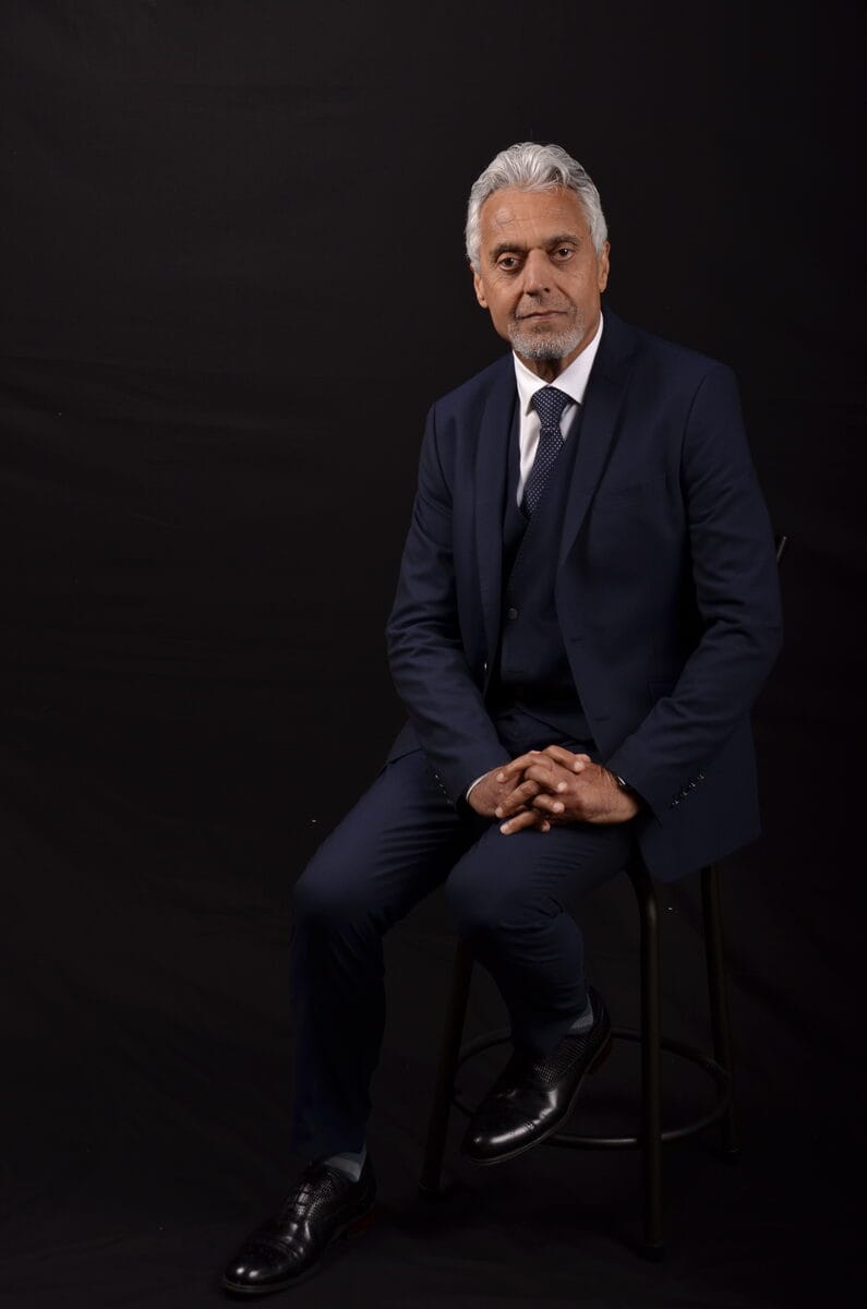 Atta Arghandiwal seated in a dark suit against a black background, in a formal editorial portrait.